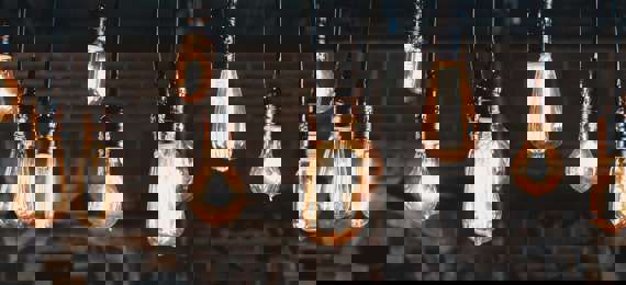 Ten different shaped light bulbs all light up and hanging from the ceiling with a brick wall in the background.  