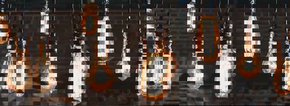 Ten different shaped light bulbs all light up and hanging from the ceiling with a brick wall in the background