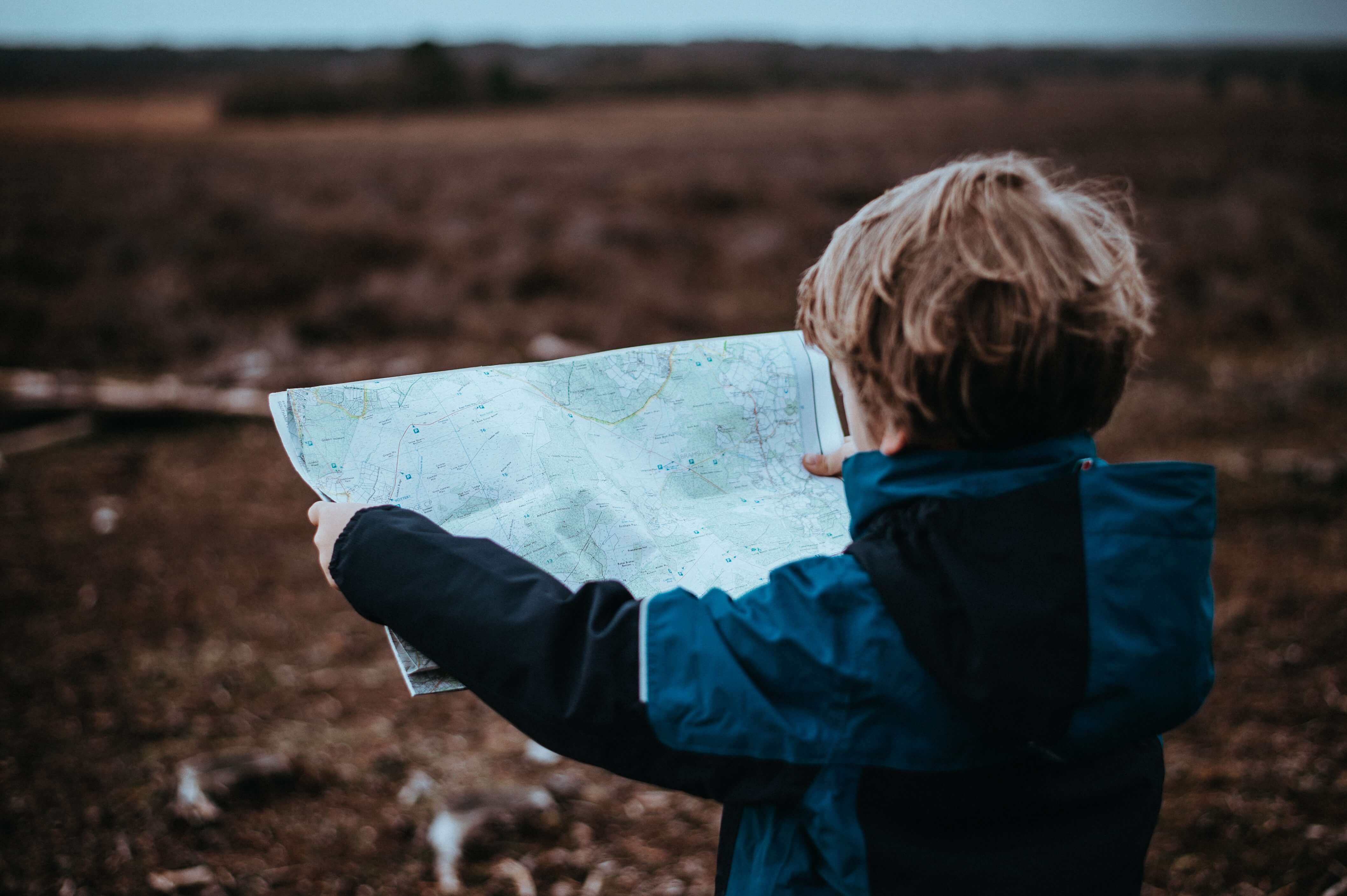 A child outside, holding a map in front of him with his back to the camera.