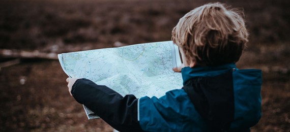 A child outside, holding a map in front of him with his back to the camera.