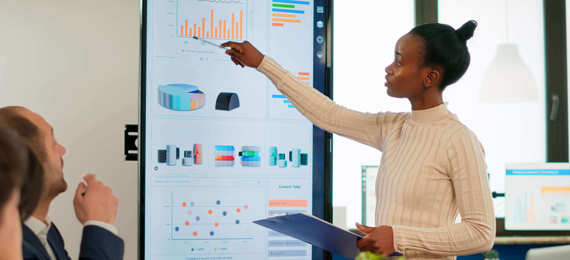 A person gives presentation to a group of people sitting at the table. The person is holding clipboard and points to the big screen showing different graphs and statistics