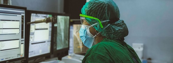 A person in a green surgical scrub wearing facemask, goggles and gloves enters information on the PC in a hospital.
