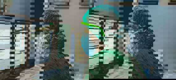 A person in a green surgical scrub wearing facemask, goggles and gloves enters information on the PC in a hospital.