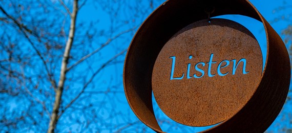 Outdoor sculpture that looks like rusted metal circle with word "listen" cut out in the middle on a blue sky background.