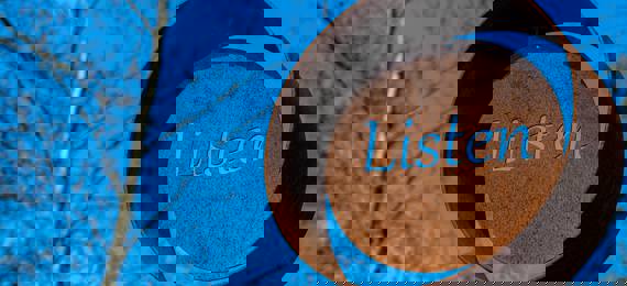 Outdoor sculpture that looks like rusted metal circle with word "listen" cut out in the middle on a blue sky background.